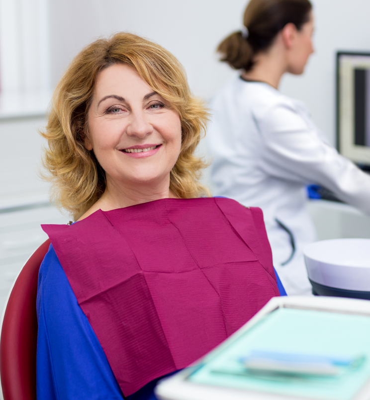 In the foreground, a middle age female patient wearing a dental bib smiles at the camera. A dental provider stands in the background.